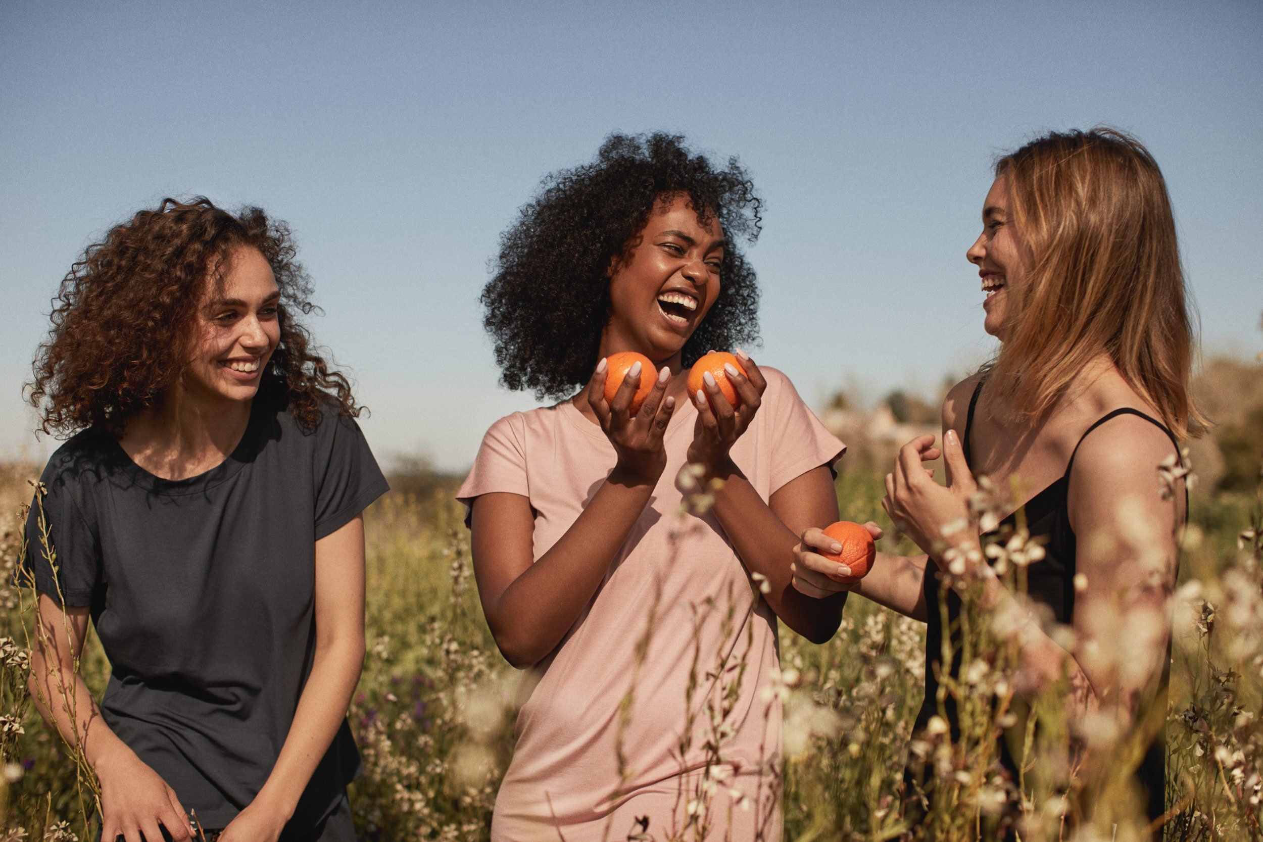 Three women laugh together in a sunny field, holding oranges. One is wearing a pink t-shirt, one is wearing a black t-shirt.