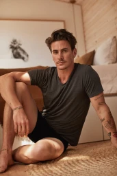 Man with tattoos seated on a jute rug in a bedroom wearing a grey t-shirt and black shorts, photographed by Petter Karlstrom.