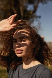 Portrait of a woman with curly brown hair raising her hand to shield her face from bright sunlight outdoors.