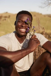 Smiling Black man holding purple wildflowers in front of his eye outdoors in a grassy rural landscape by photographer Petter Karlstrom