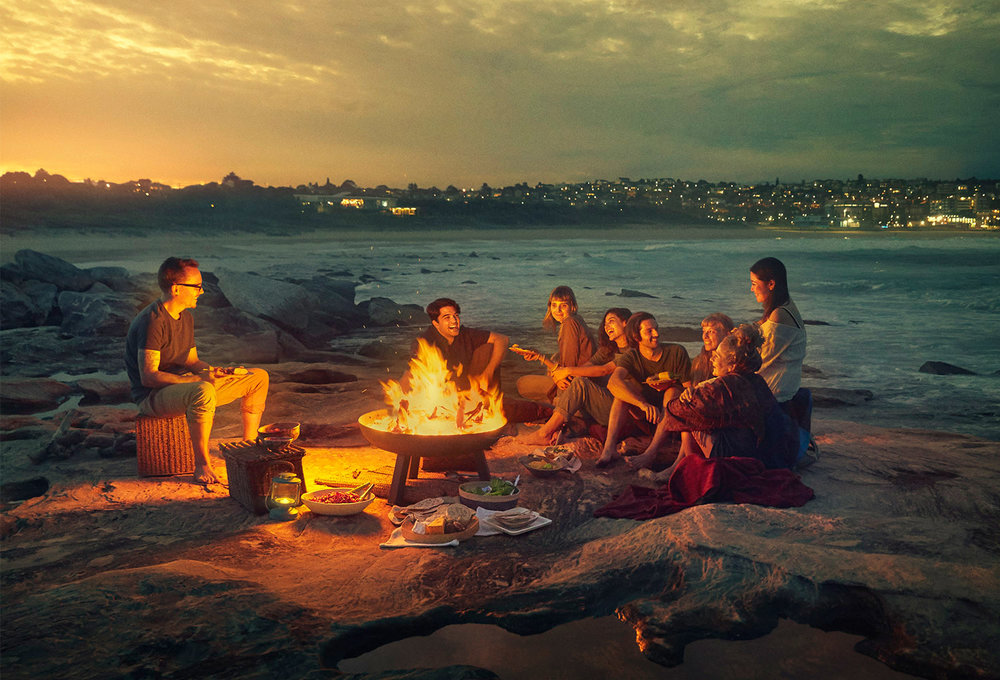 Group of people gathered around a fire pit on a rocky beach at dusk, with city lights visible in the background, enjoying food and music.