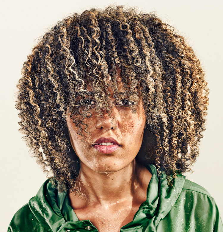 Portrait of an adult woman with curly hair, wearing a green, hooded raincoat in front of a neutral background.
