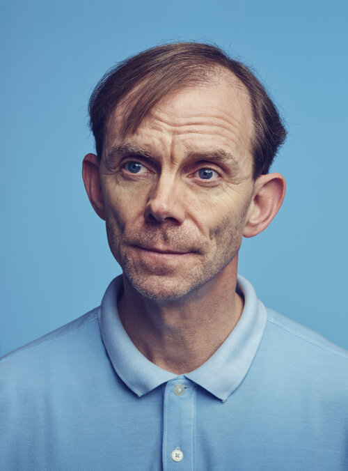Headshot of a mature adult male with thinning brown hair, wearing a light blue polo shirt, against a light blue background.