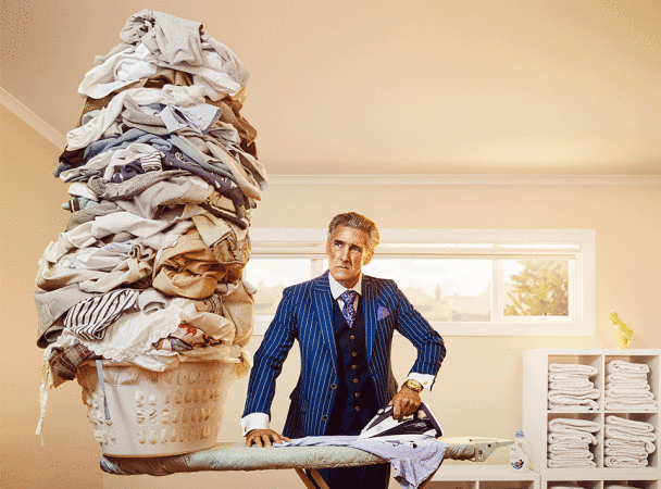 Mature man in suit ironing laundry near large laundry pile. The scene is set indoors in a well-lit room with natural light.