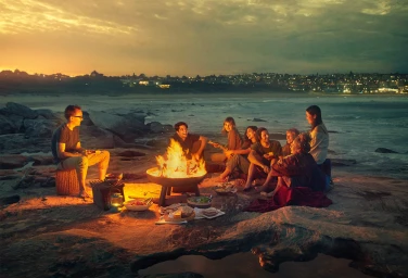 Group of diverse people gathered around a glowing bonfire on a rocky beach at dusk with distant town lights visible