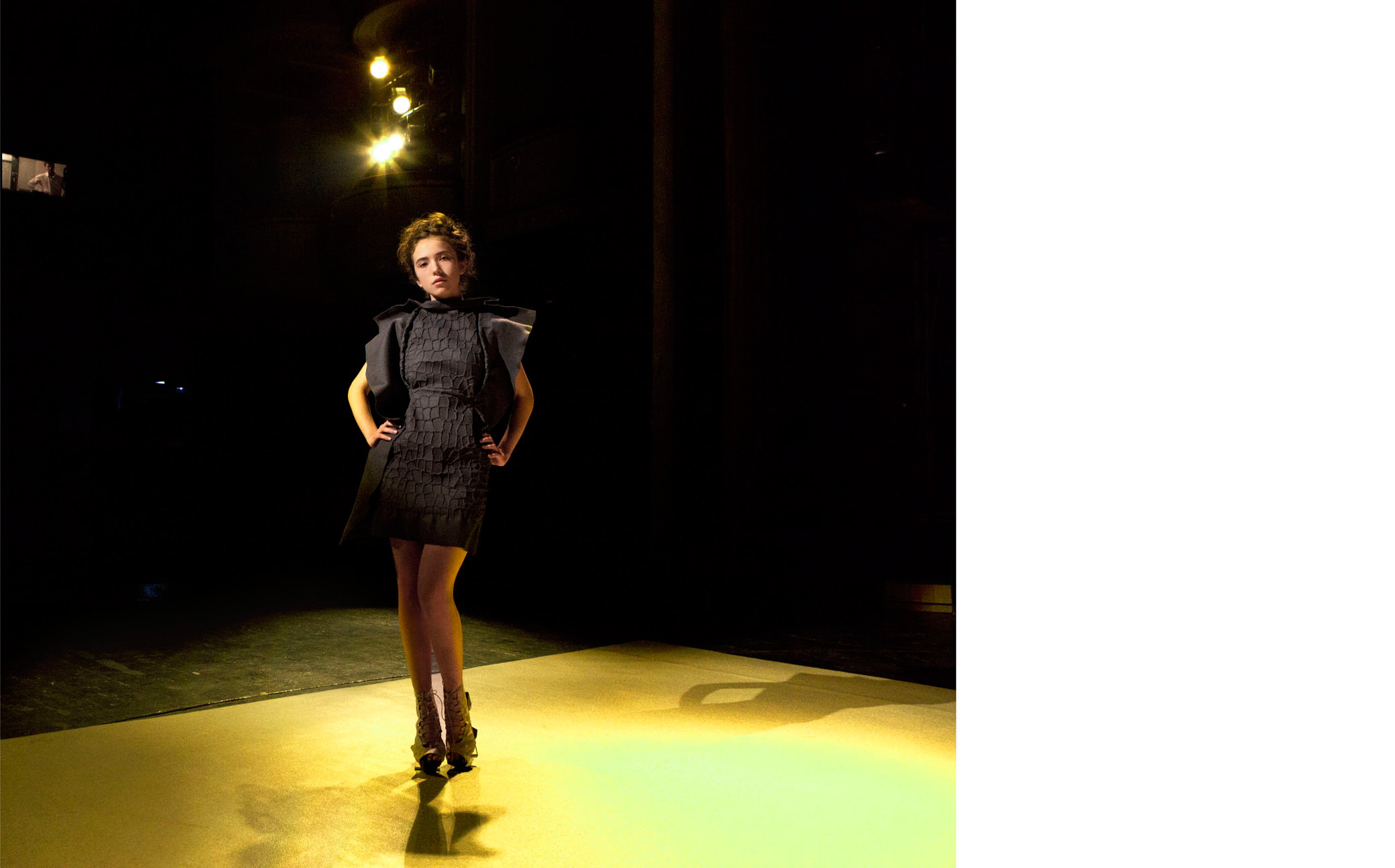 Fashion runway shot of a female adult in a gray dress and heeled shoes with her hands on her hips on a yellow-lit runway stage.