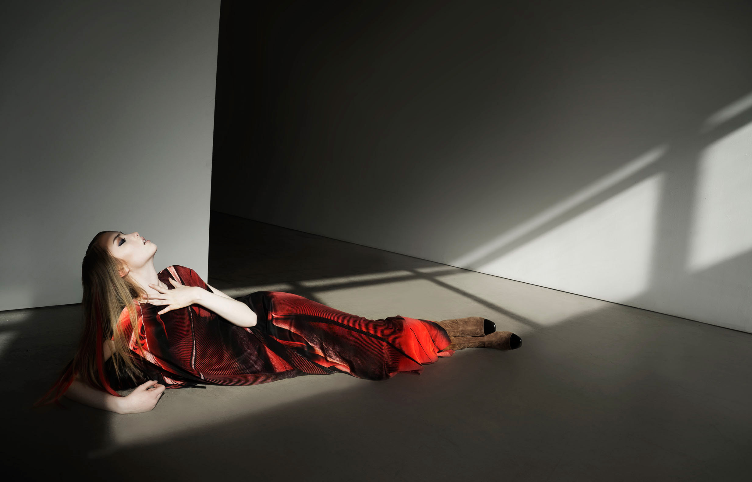 Woman in a red dress lies on a gray floor, eyes looking upward, with light and shadows creating contrast on the walls and floor.