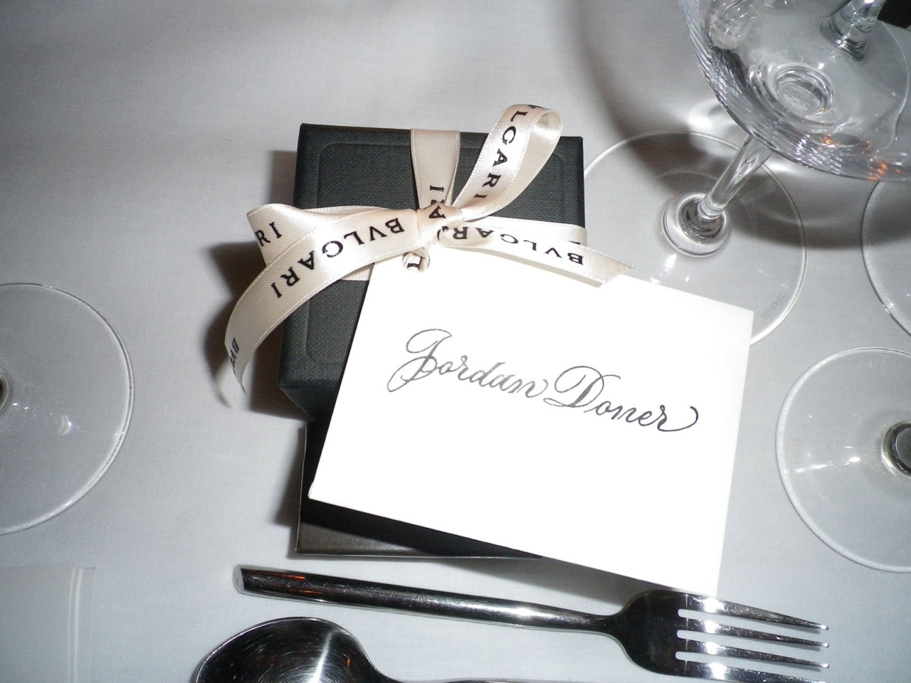 Close-up of a black gift box tied with a Bulgari ribbon and a card that reads 'Jordan Doner,' alongside silverware and wine glasses on a white table.