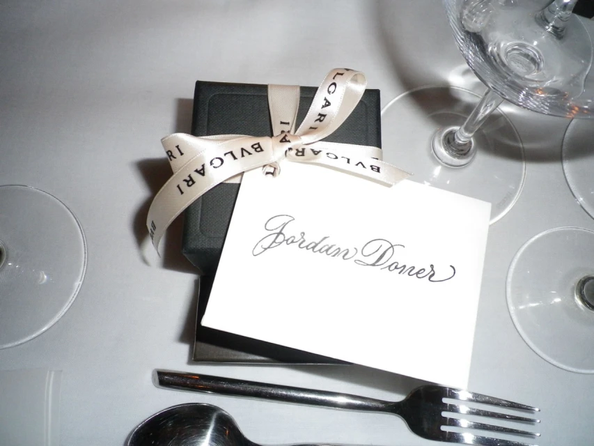 Close-up of a black gift box tied with a Bulgari ribbon and a card that reads 'Jordan Doner,' alongside silverware and wine glasses on a white table.