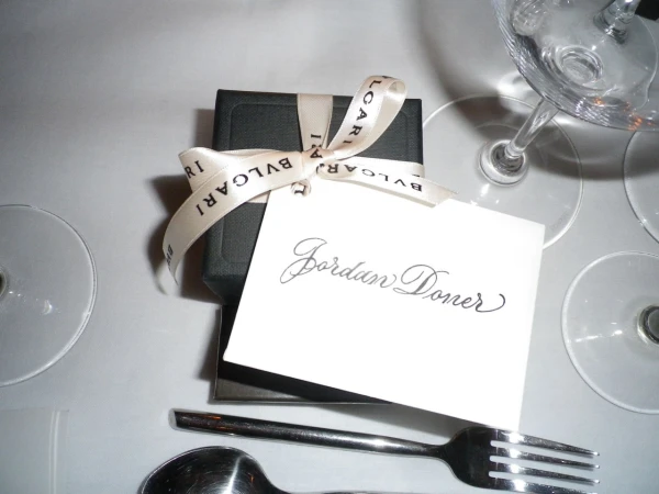 Close-up of a black gift box tied with a Bulgari ribbon and a card that reads 'Jordan Doner,' alongside silverware and wine glasses on a white table.