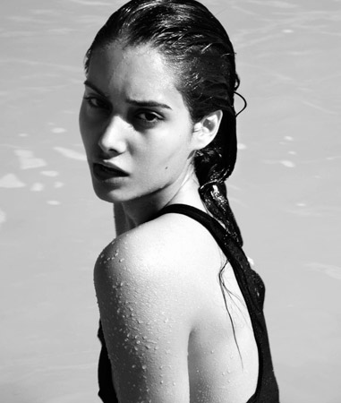 Black and white portrait of a young woman with wet hair, wearing a dark top, and looking over her shoulder at the camera outdoors.