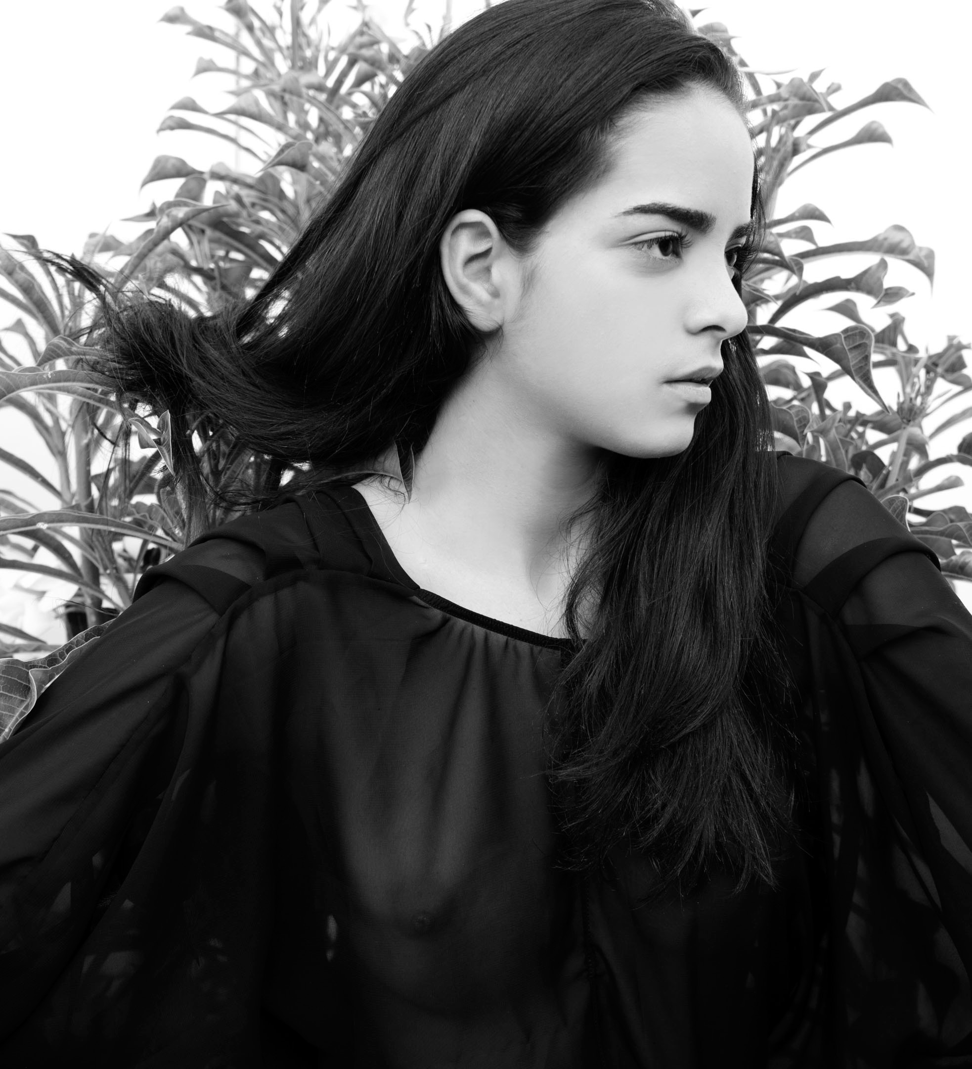 Black and white portrait of a young woman with long, dark hair wearing a sheer black top, posing in front of green plants.