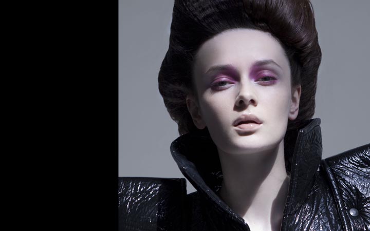 Portrait of a model with dramatic dark hair and heavy pink makeup. She wears a glossy, high-necked black vinyl top in a studio shot.