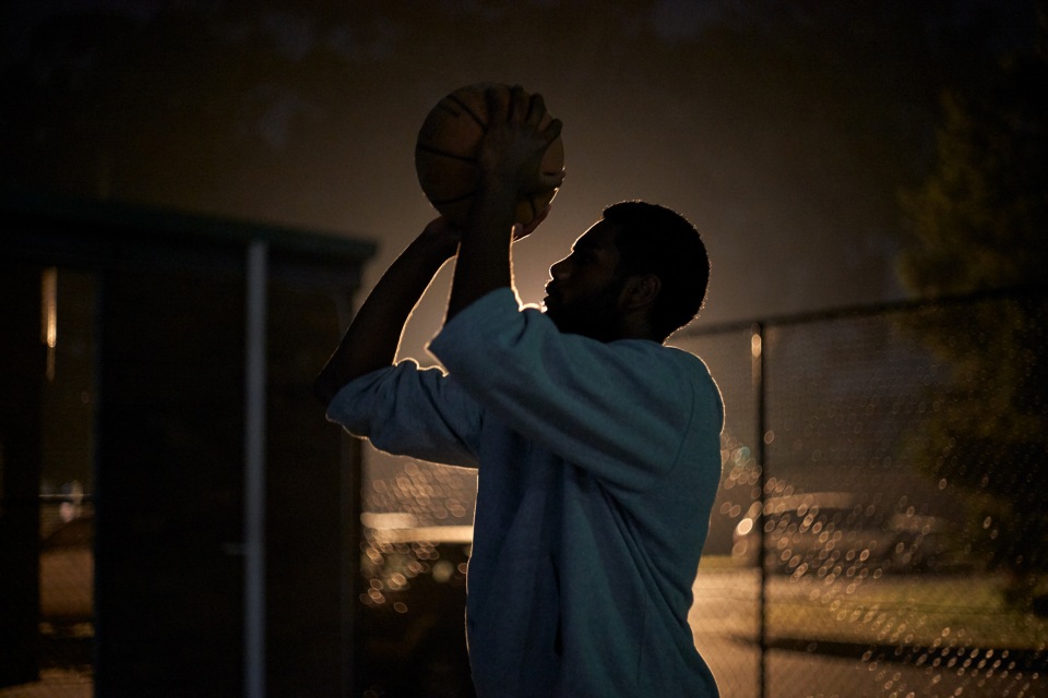 Silhouette of an African-American man in a white sweatshirt shooting a basketball outdoors at night; chainlink fence and trees in background.