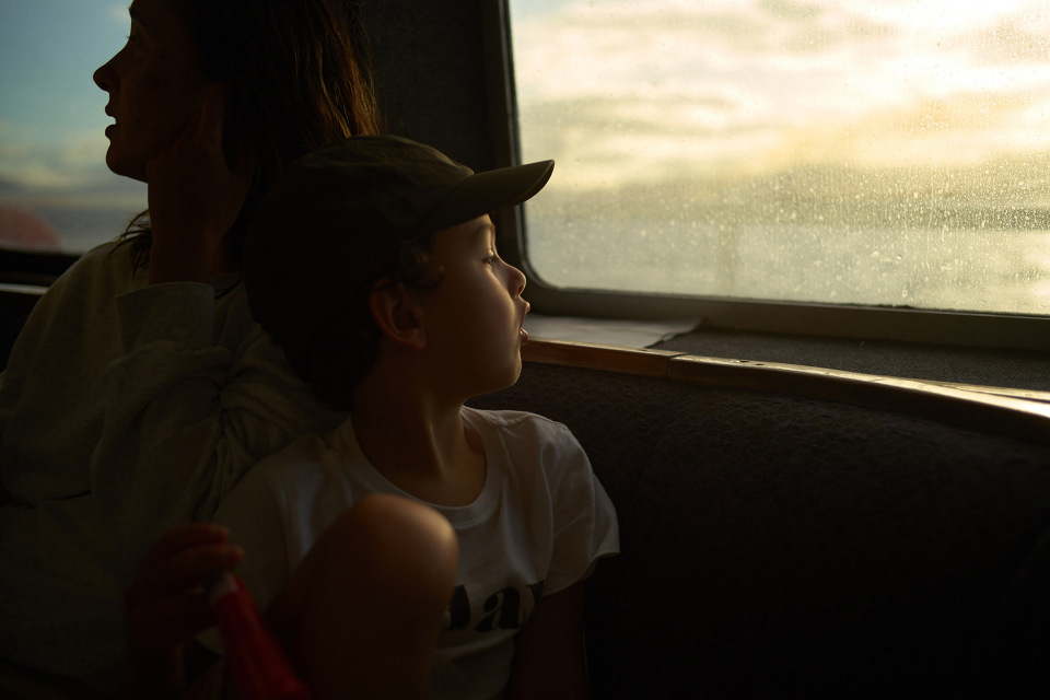 Child in cap looks out of a window on a boat with a woman behind him. Window shows a bright seascape scene, backlighting the family.
