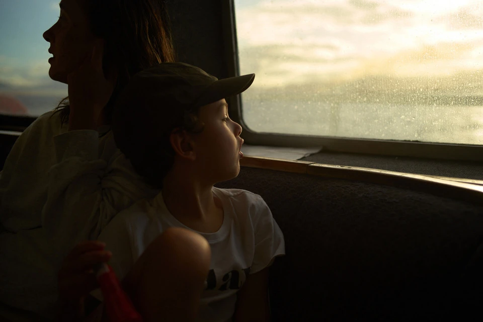 Child in cap looks out of a window on a boat with a woman behind him. Window shows a bright seascape scene, backlighting the family.