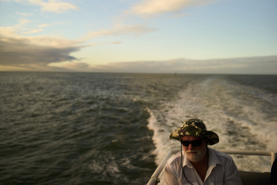 Man with white beard and camo hat rides boat in ocean, sunny day, visible wake. Blue and white sky above the water.
