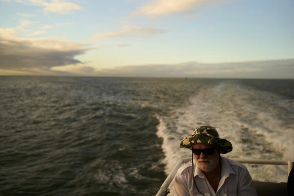 Man with white beard and camo hat rides boat in ocean, sunny day, visible wake. Blue and white sky above the water.