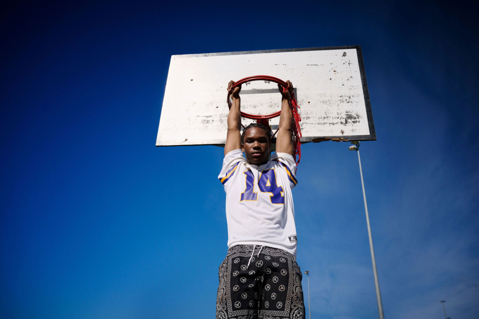 Teenage boy in sports jersey hangs from basketball hoop against a blue sky on a sunny day in an outdoor court setting.