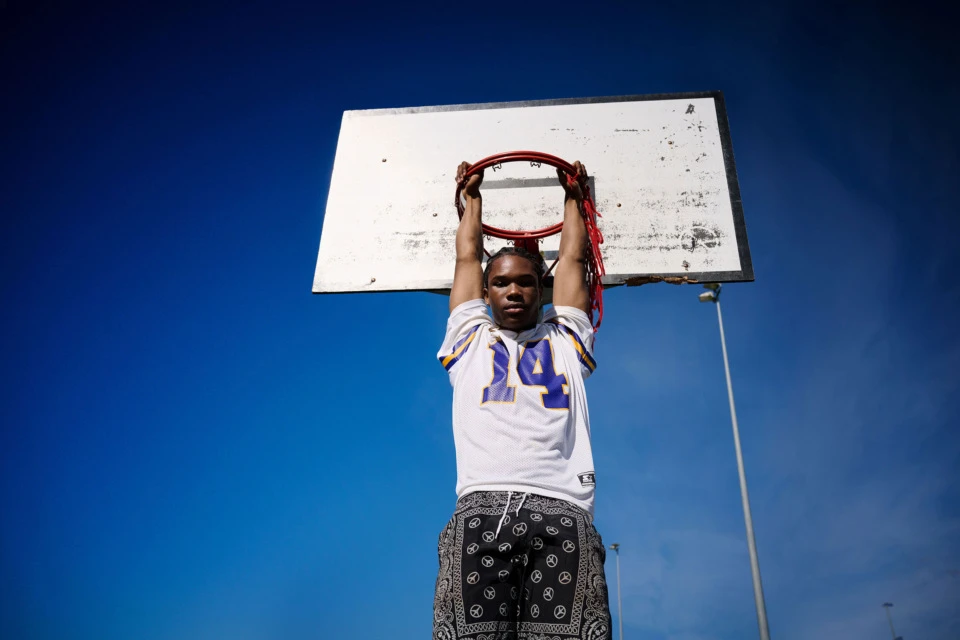 Teenage boy in sports jersey hangs from basketball hoop against a blue sky on a sunny day in an outdoor court setting.