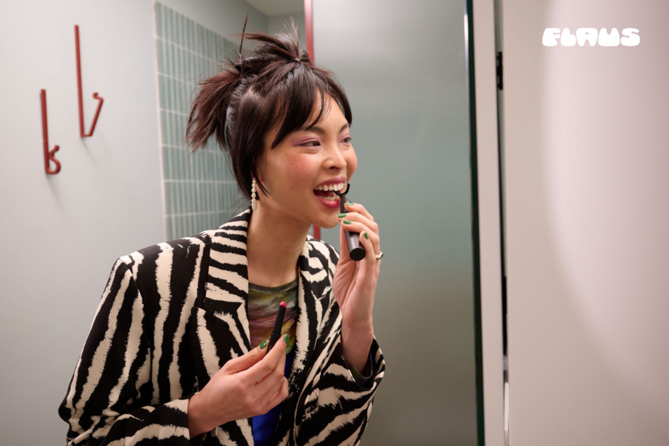 Smiling woman in zebra-print blazer applies lip makeup in a bright, modern bathroom setting near white door and wall-mounted hooks.