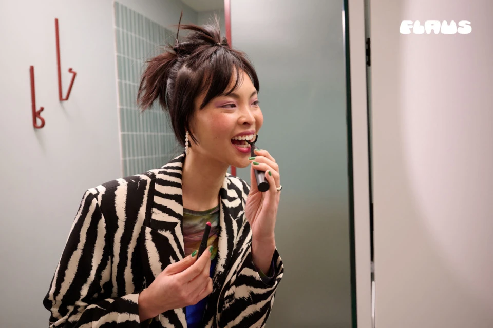 Smiling woman in zebra-print blazer applies lip makeup in a bright, modern bathroom setting near white door and wall-mounted hooks.