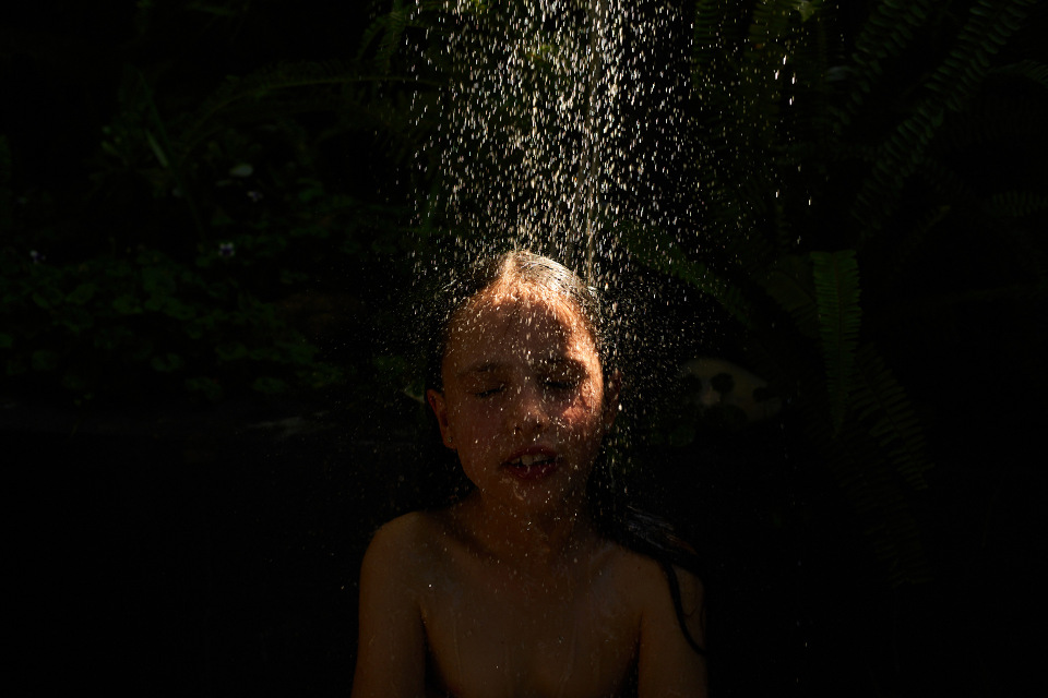Close-up portrait of a person standing under a stream of falling water droplets with eyes closed against a dark background
