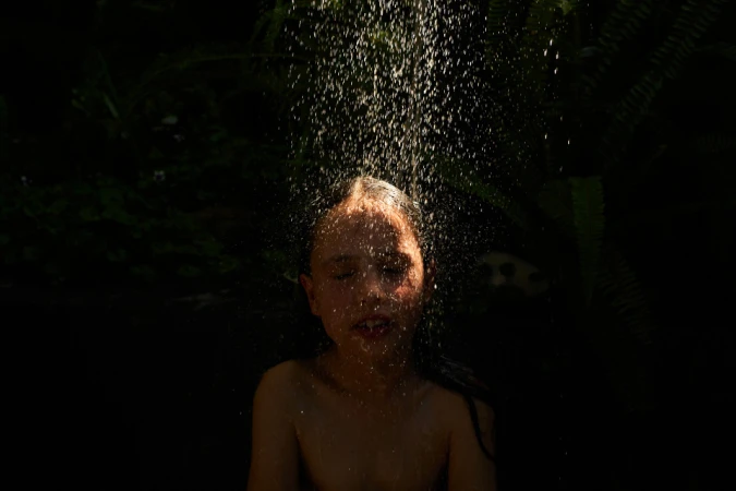 Close-up portrait of a person standing under a stream of falling water droplets with eyes closed against a dark background