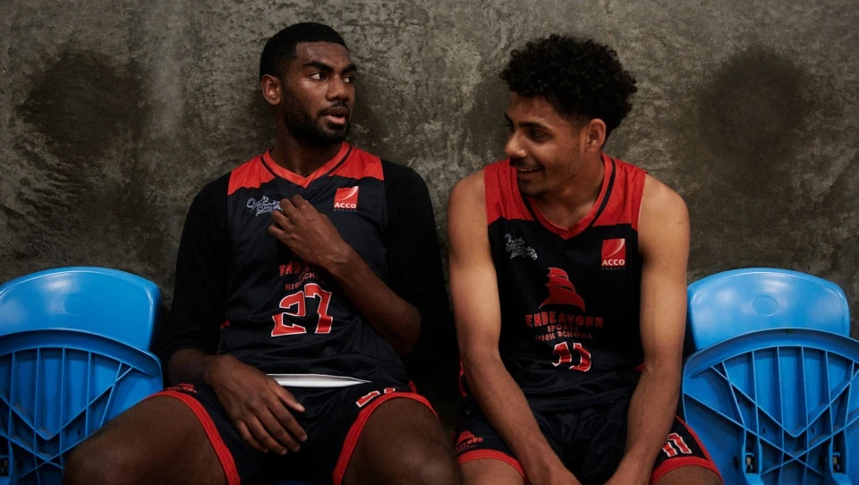 Two athletic men in jerseys sit side-by-side in an indoor setting, captured in a medium shot against a textured gray wall background.