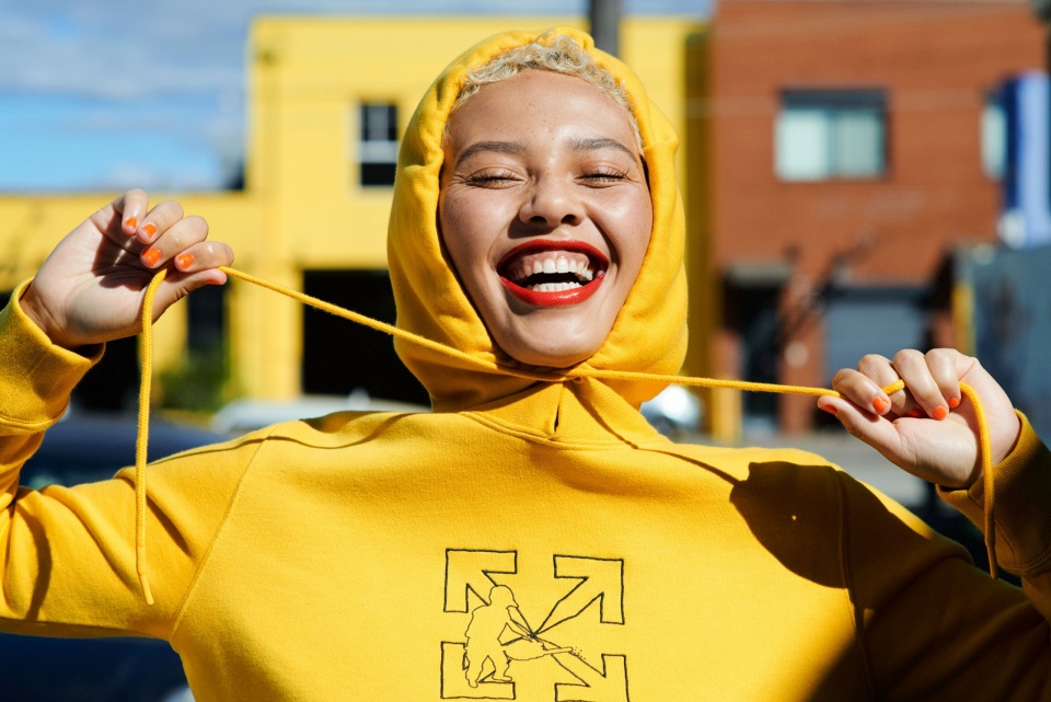 Close-up of a smiling woman in a yellow hoodie with orange nail polish, posing outdoors against colorful urban buildings.