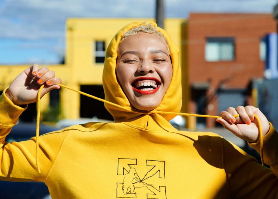 Woman with fair skin smiles, wearing a yellow hooded sweatshirt with black graphic, standing outdoors with blurred buildings behind her.