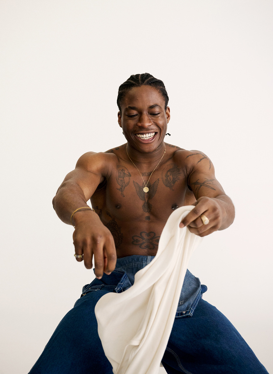 Portrait of smiling shirtless Black man with braided hair and tattoos, holding white folded shirt, wearing jeans and jewelry against white backdrop.