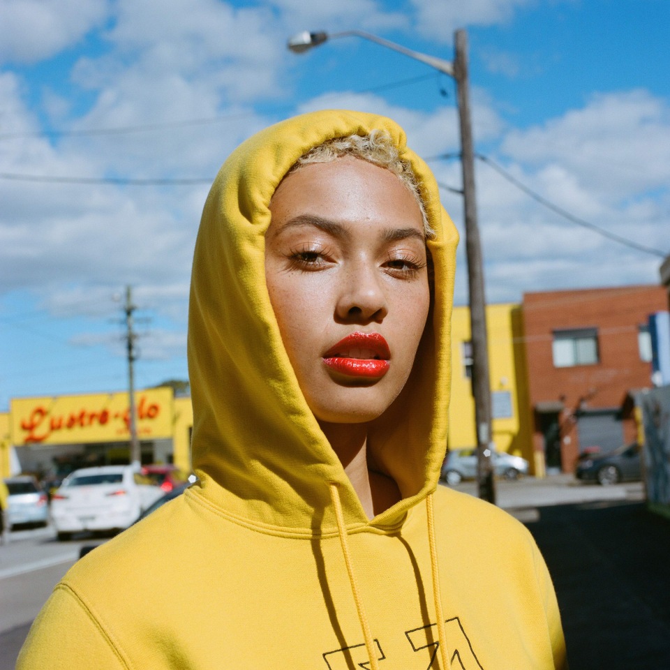 Portrait of a young adult woman with short blonde hair wearing a yellow hooded sweatshirt, red lipstick, and standing outdoors in daytime.