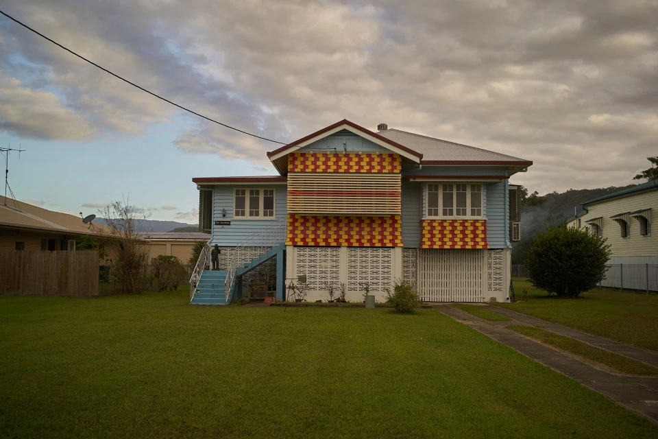 Two-story light blue house with red and yellow awning details on a green lawn under a cloudy sky, viewed from a distance.