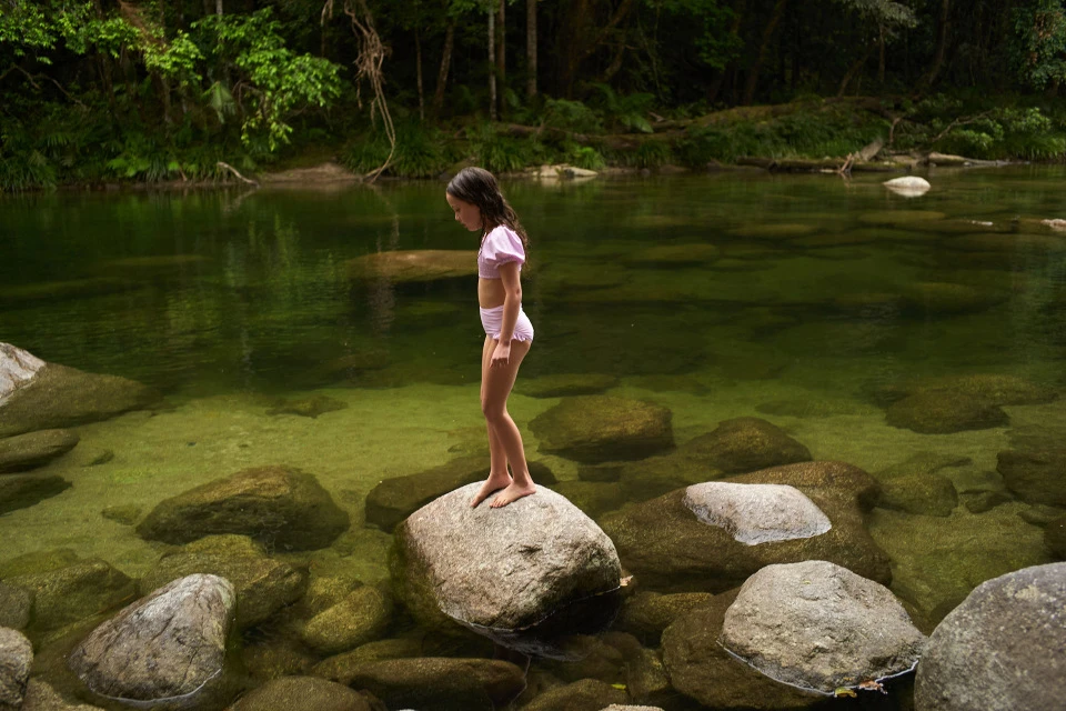 A child in a pink swimsuit stands on a rock in a clear stream surrounded by lush greenery, reflecting the natural light in a tranquil scene.