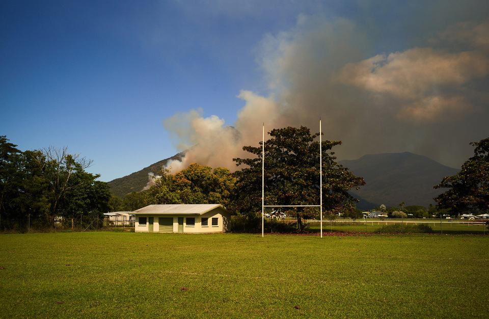 Landscape view with a green field, trees, mountains, smoke, and a clear blue sky with white clouds. An outdoor sports goal is visible.