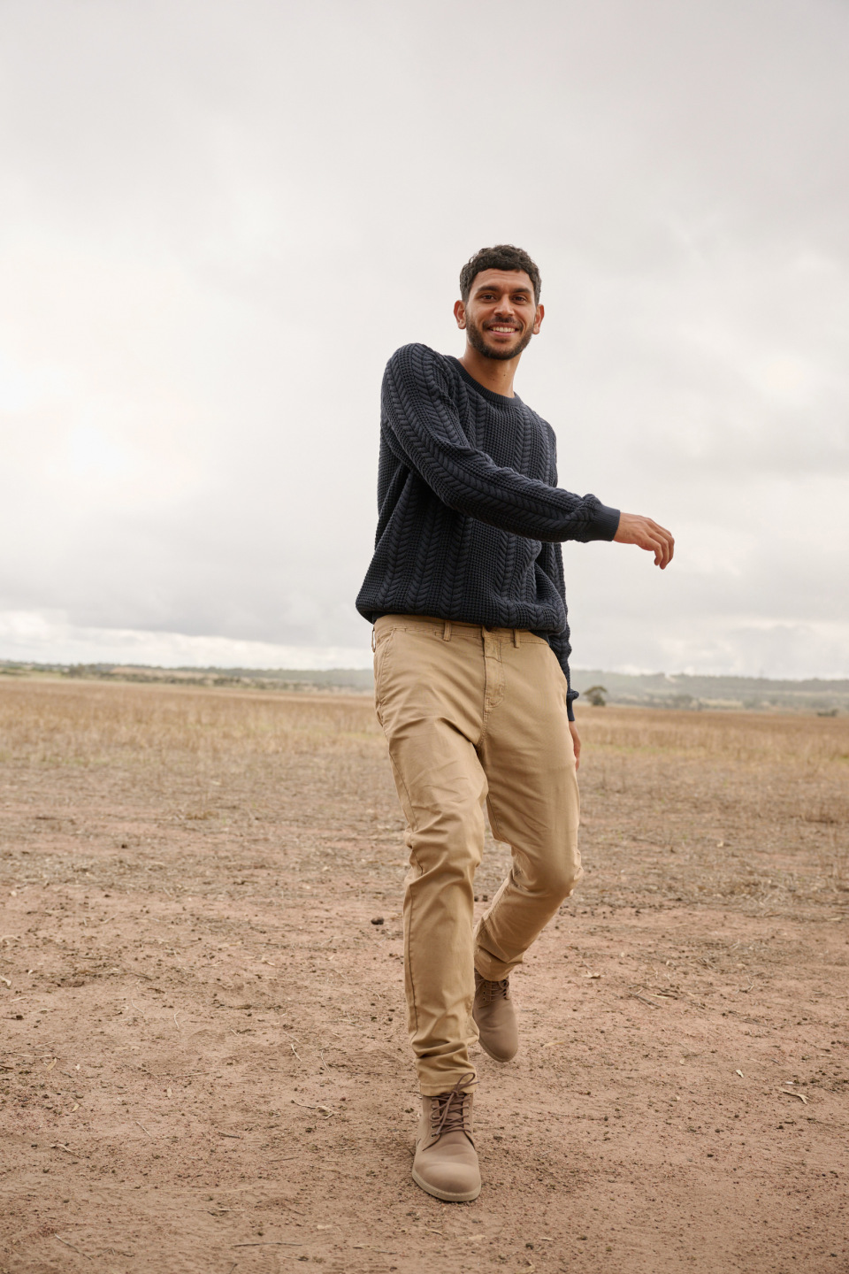 A young adult man walks across a field wearing a sweater, tan pants and boots against a cloudy sky in a medium outdoor shot.