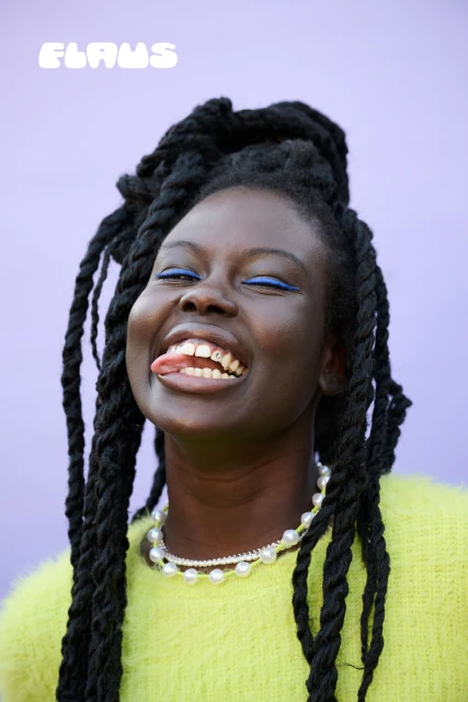 Smiling Black woman with braided hair, pearl necklace, and bright green top against a lavender background, sticking out her tongue.