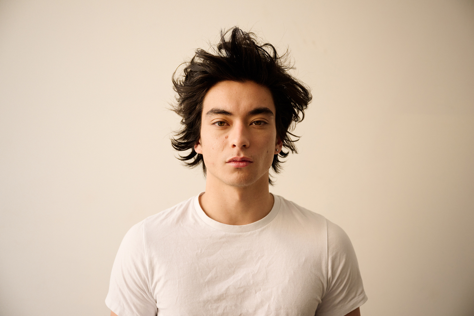 Portrait of a young man with dark tousled hair wearing a white t-shirt against a plain light wall