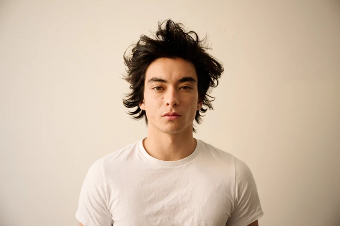 Portrait of a young man with dark tousled hair wearing a white t-shirt against a plain light wall