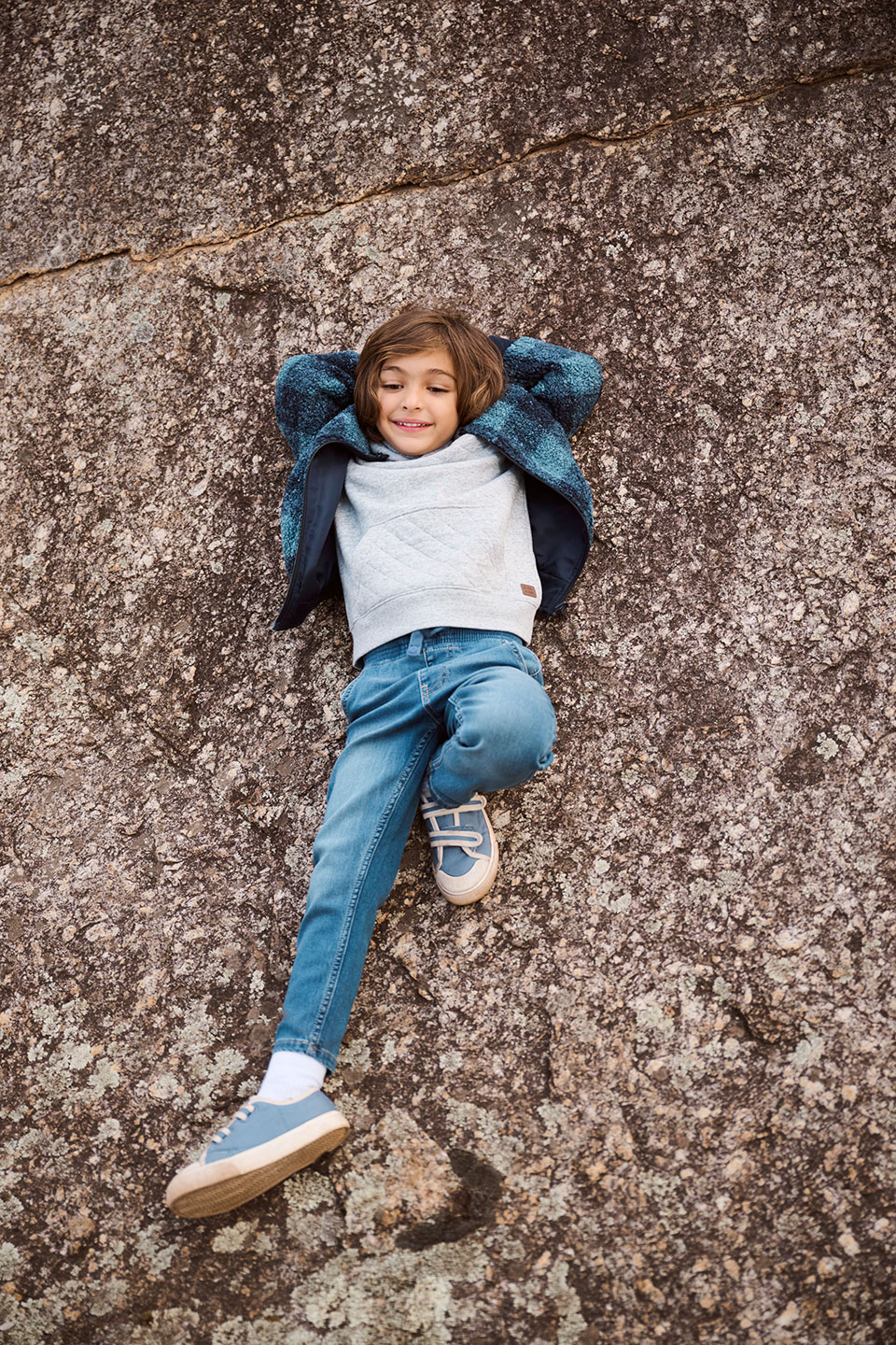 Young child lays on a large rock with hands behind head wearing a blue jacket, gray sweater, jeans, and blue shoes while smiling.