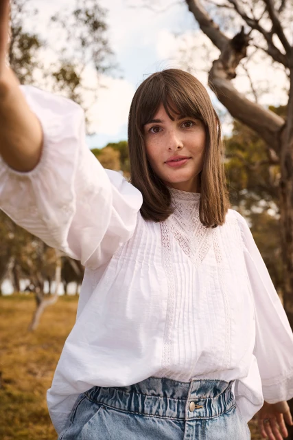 A woman with brown hair wears a white peasant blouse and high-waisted denim jeans, standing outdoors with trees and sunlight in the background.