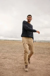 Smiling man walking outdoors in a textured dark sweater and khaki trousers under an overcast sky.