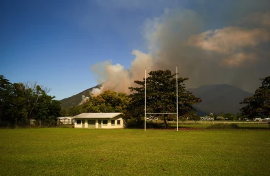 A rugby goalpost stands on a green sports field with a small building, trees, and dark smoke rising from a mountain behind it.