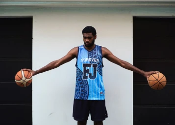 Black man holding two basketballs out to the sides while wearing a blue patterned basketball jersey and dark shorts
