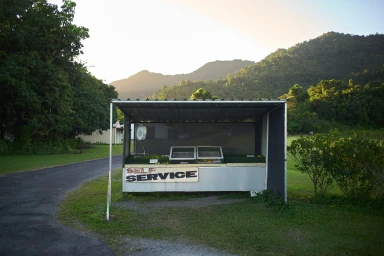 A roadside self-service fruit stand under a corrugated metal shelter with mountain and forest background in Sydney by Tobias Rowles.