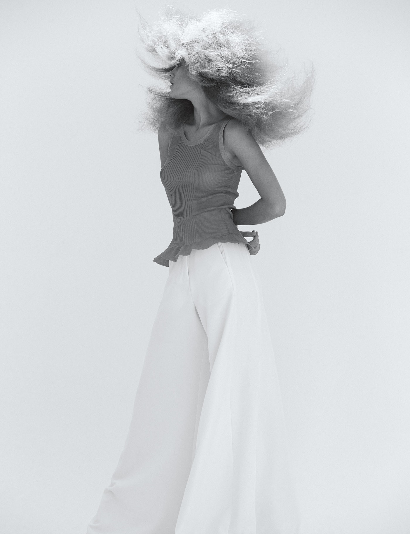 Woman with voluminous blonde hair wearing a grey top and white pants poses in front of a white background in a studio shot.