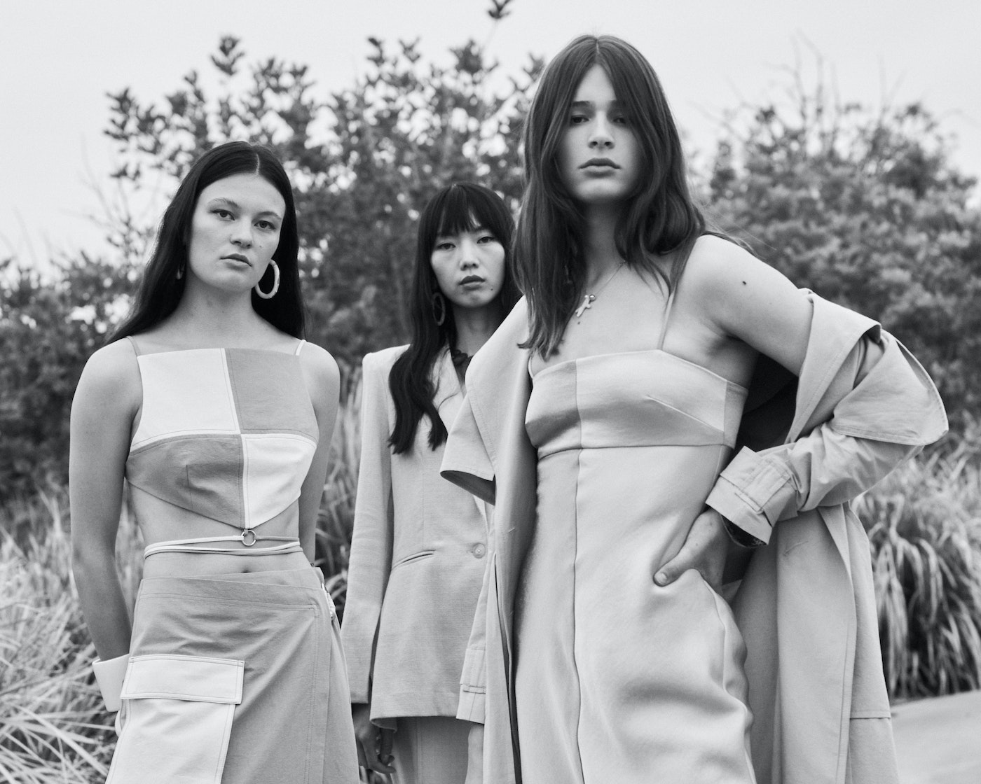 Three female fashion models in earth-tone dresses pose in an outdoor setting with greenery and an overcast sky in a black-and-white photo.