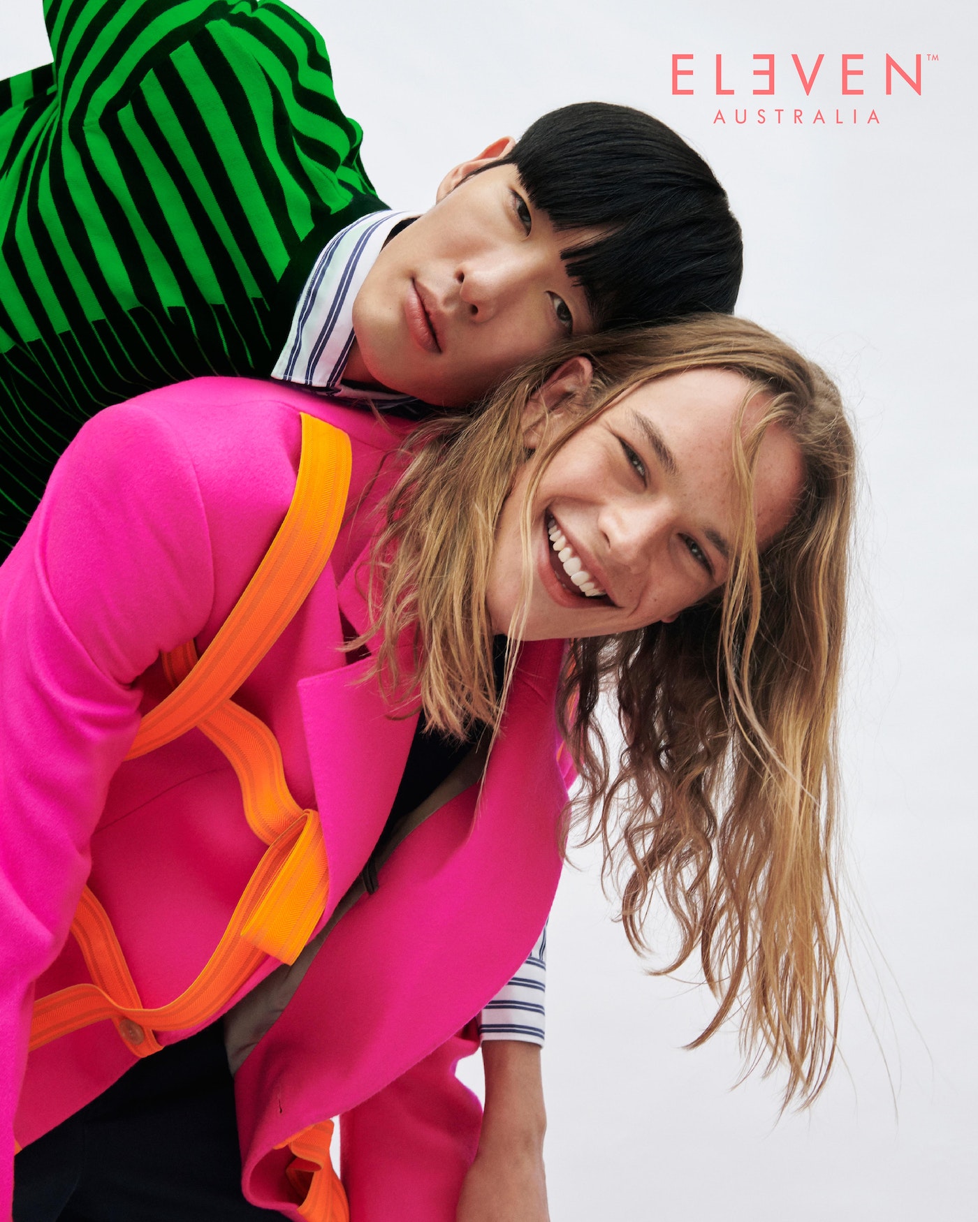 Two young adults in brightly colored blazers pose in a light studio. One smiles openly, showing teeth, with Eleven Australia logo in corner.