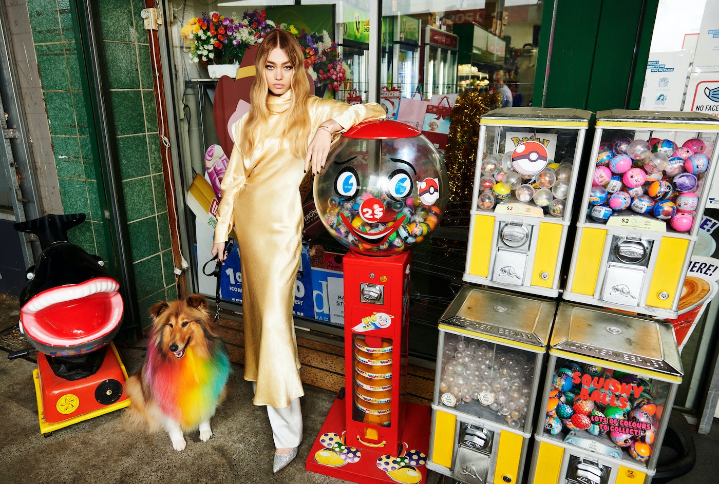 Woman in gold dress with dog next to gumball machines and vintage toy ride in front of store, fashion editorial photography.